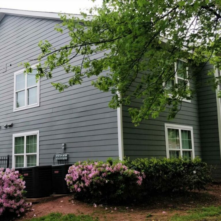 close up view of the corner of an apartment with gray siding surrounded by trees and bushes