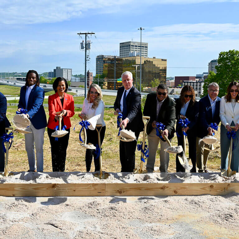 A diverse group of city leaders and community members breaking ground at a new development site in Chattanooga’s Westside, holding shovels with blue ribbon bows, against the backdrop of a city skyline and highway.