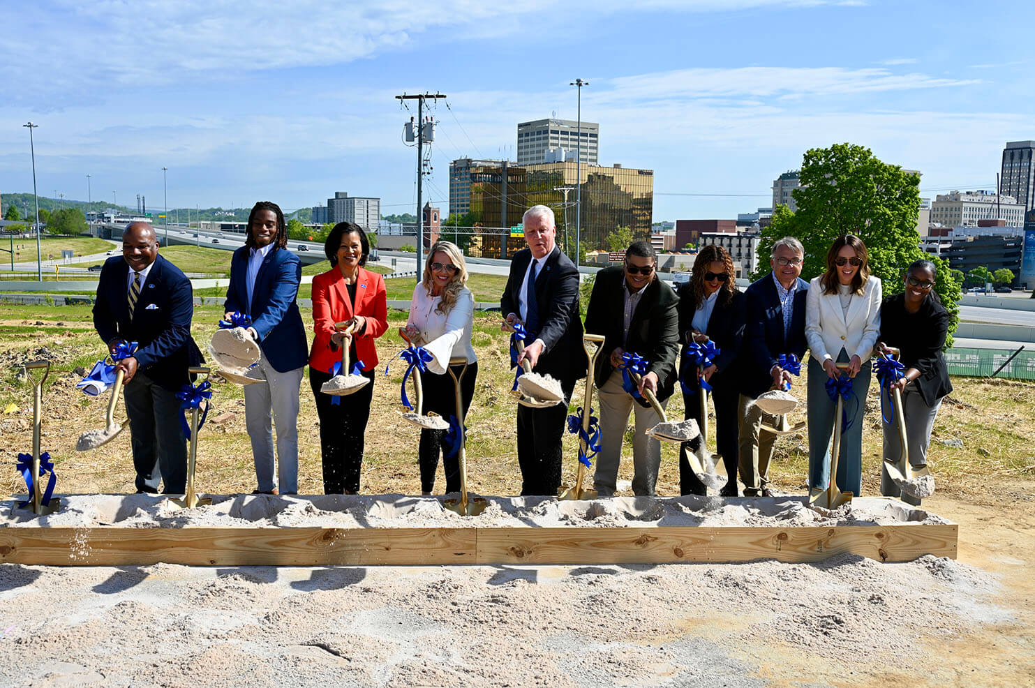 A diverse group of city leaders and community members breaking ground at a new development site in Chattanooga’s Westside, holding shovels with blue ribbon bows, against the backdrop of a city skyline and highway.