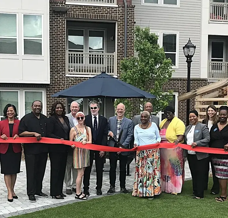 A diverse group of people stands in front of a new apartment building during a ribbon cutting for an Athens affordable housing complex in Georgia. The group holds a long red ribbon stretched across a landscaped courtyard with green grass and a blue umbrella in the background. The building behind them features modern brick and siding with balconies and large windows, symbolizing the grand opening of a new residential community.