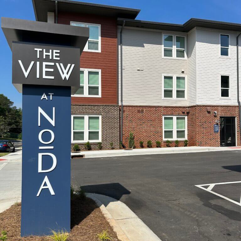 Exterior view of The View at NoDa apartment complex on a clear sunny day, featuring a modern three-story building with brick and siding facade and a large dark blue monument sign in the foreground with white text reading 'The View at NoDa.