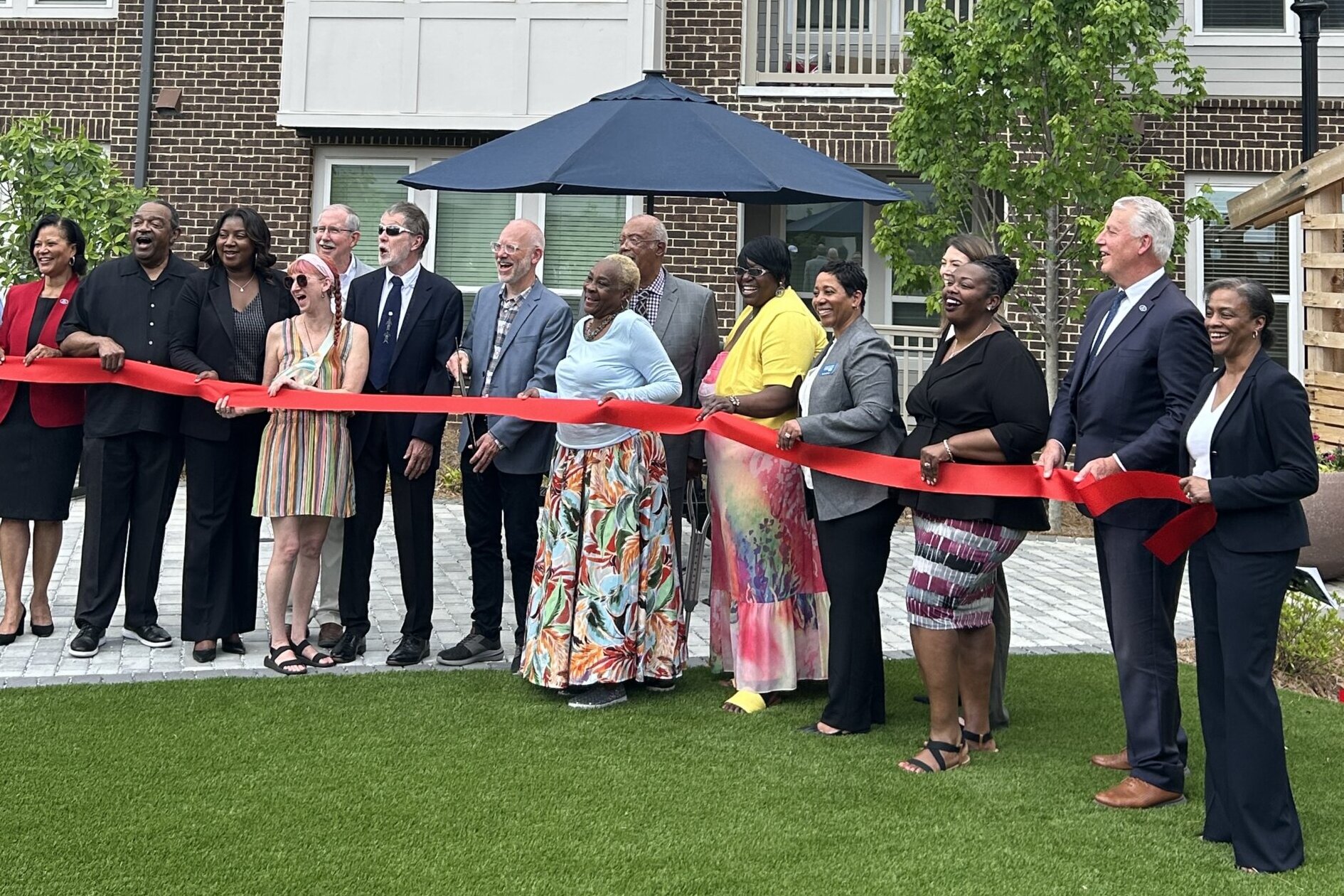 A group of people stands in a line holding a red ribbon at a ribbon-cutting ceremony in front of a new apartment building. The group includes men and women of diverse backgrounds, dressed in both business and casual attire, smiling in celebration. Behind them is a modern, multi-story building with brick, white, and red siding, along with a blue patio umbrella and landscaped greenery.