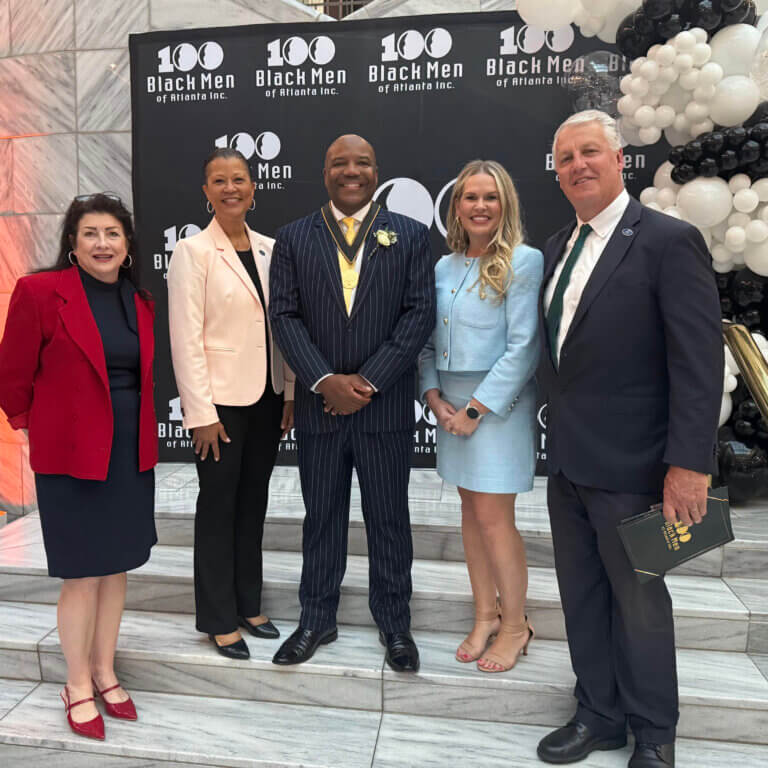 Five professionally dressed individuals stand on marble stairs in front of a black backdrop with the logo "100 Black Men of Atlanta Inc." in white. The group includes three women and two men, smiling for the camera. One man in the center wears a dark pinstriped suit and gold medallion. Black and white balloon decorations are visible.
