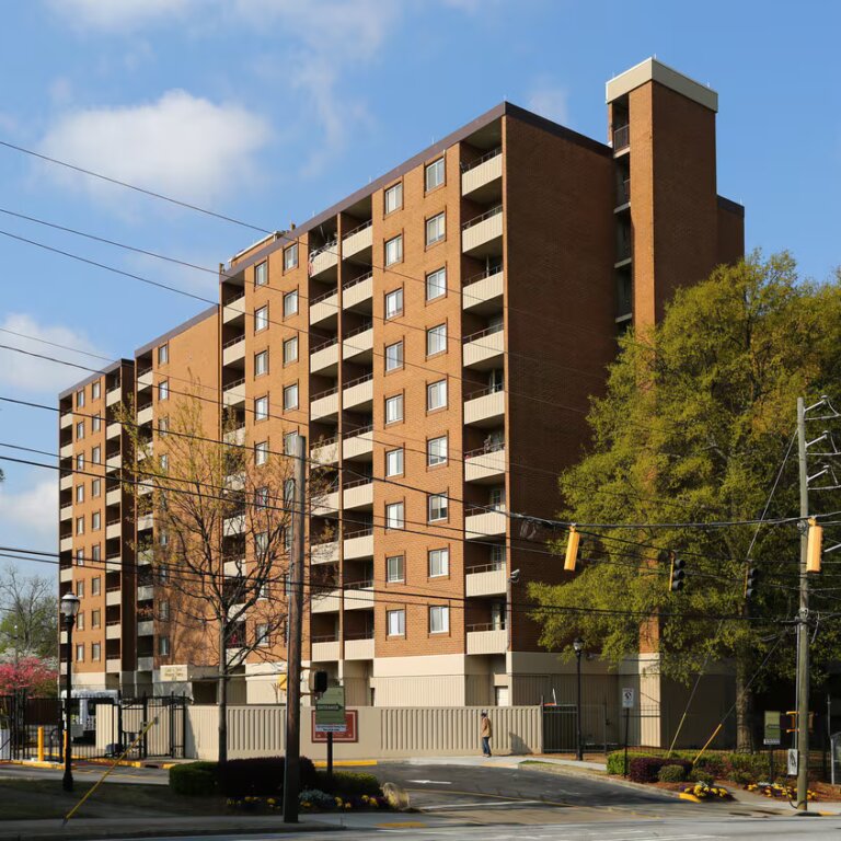 Exterior view of Cosby Spear Highrise, a multi-story senior residential building in Atlanta, Georgia, featuring a red brick facade with rows of windows and surrounded by trees under a clear blue sky.