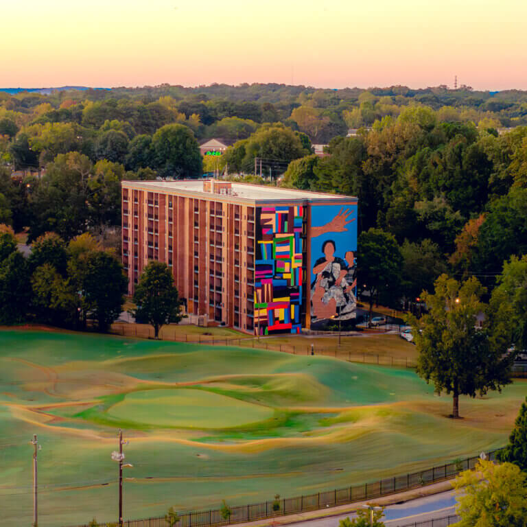 Aerial view of a mid-rise brick residential building featuring a large, colorful mural at Legacy at East Lake on its side, surrounded by trees and a nearby golf course under a warm sunset sky.