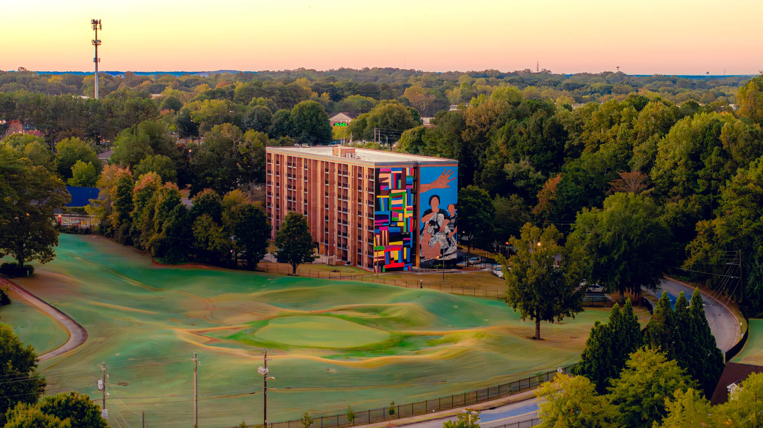 Aerial view of a mid-rise brick residential building featuring a large, colorful mural at Legacy at East Lake on its side, surrounded by trees and a nearby golf course under a warm sunset sky.