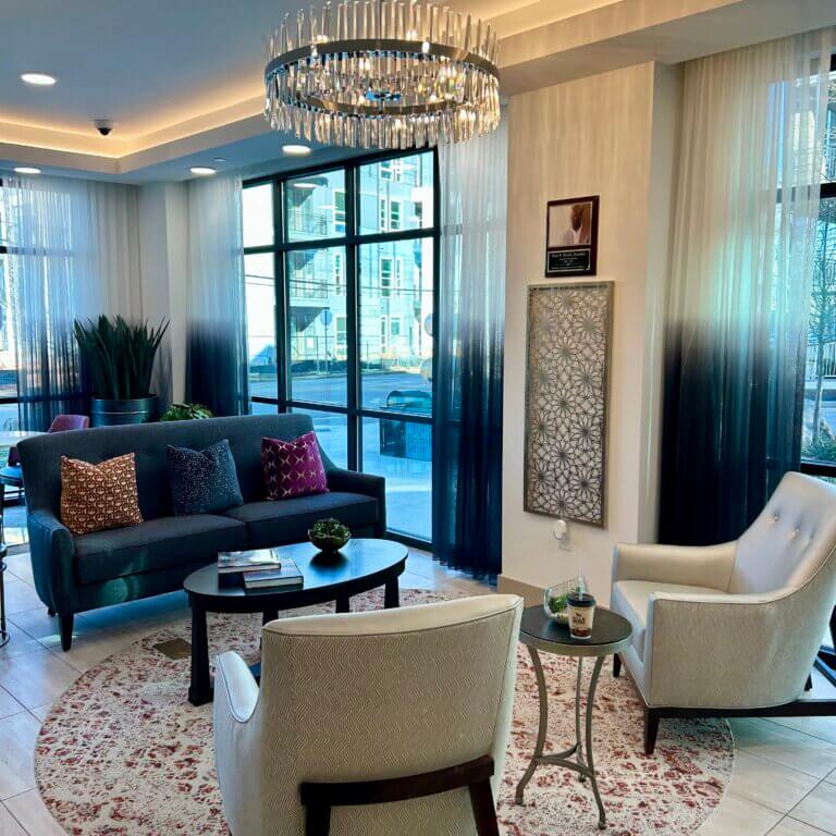 Modern lounge area at The Link featuring a gray sofa with accent pillows, two upholstered armchairs, a round coffee table, decorative rug, large windows with sheer curtains, and a contemporary chandelier overhead.