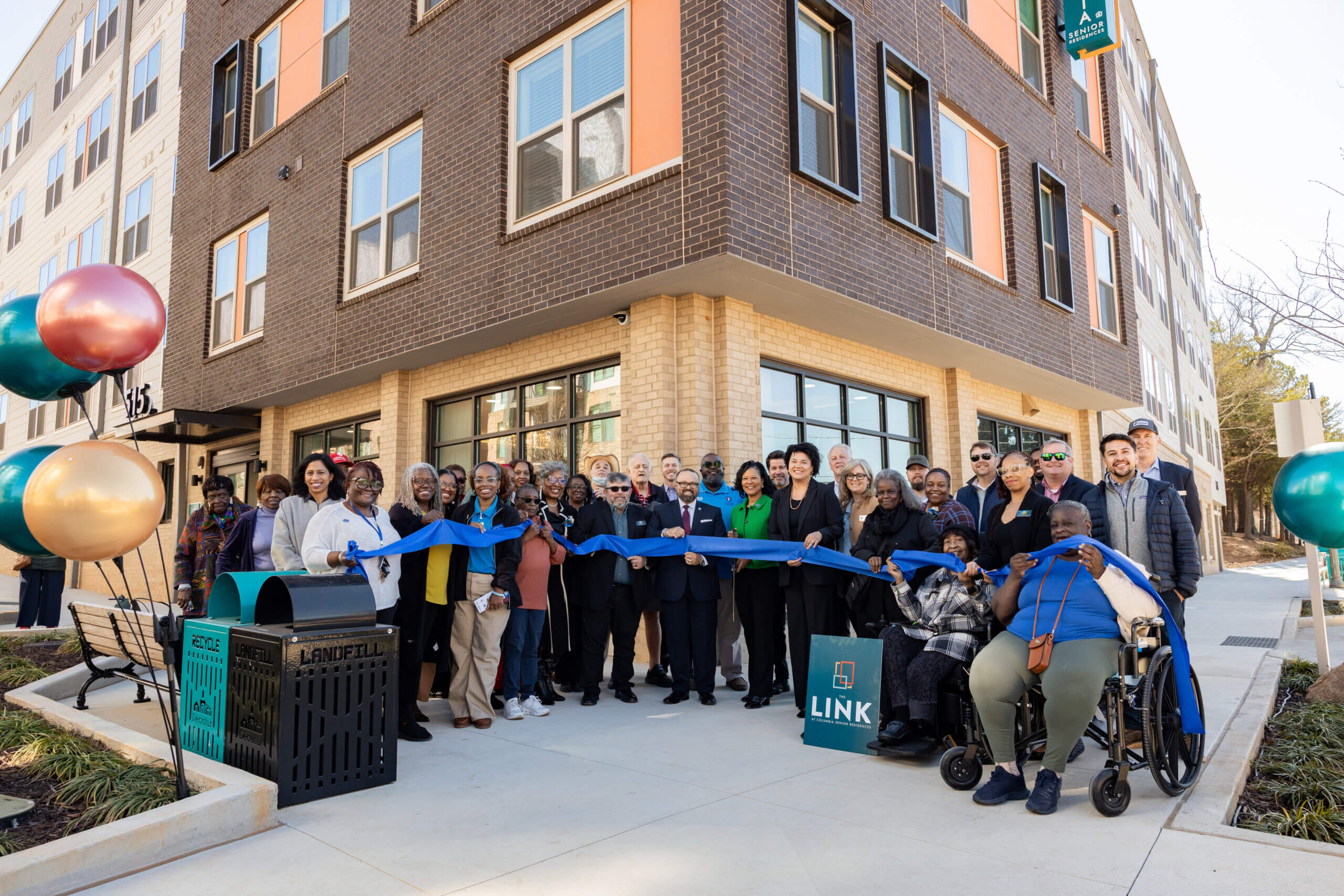 Residents, community leaders, and partners gather outside The Link at MLK for a ribbon-cutting ceremony, holding a blue ribbon in front of the newly opened residential building.