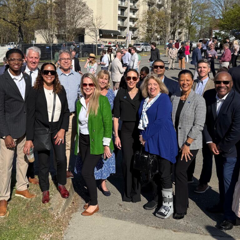 At the groundbreaking for Phase 2 of One Westside, a group of people standing outside, smiling at the camera. Trees, building, and a construction fence are visible in the background.