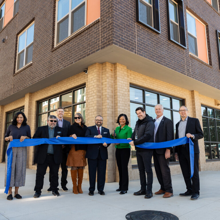 A group of community leaders and project partners stand outside the newly constructed apartment building at a senior housing development in Decatur — The Link. These individuals are holding a long blue ribbon during a ribbon-cutting ceremony. The modern mixed-use building features brick and large windows, with ground-floor retail space and residential units above, marking the opening of a new housing community.