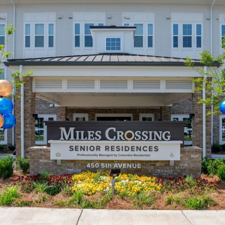 Entrance sign for Miles Crossing Senior Residences in front of a multi-story apartment building, with landscaped flowers and blue and gold balloons on either side, marking a celebratory event.