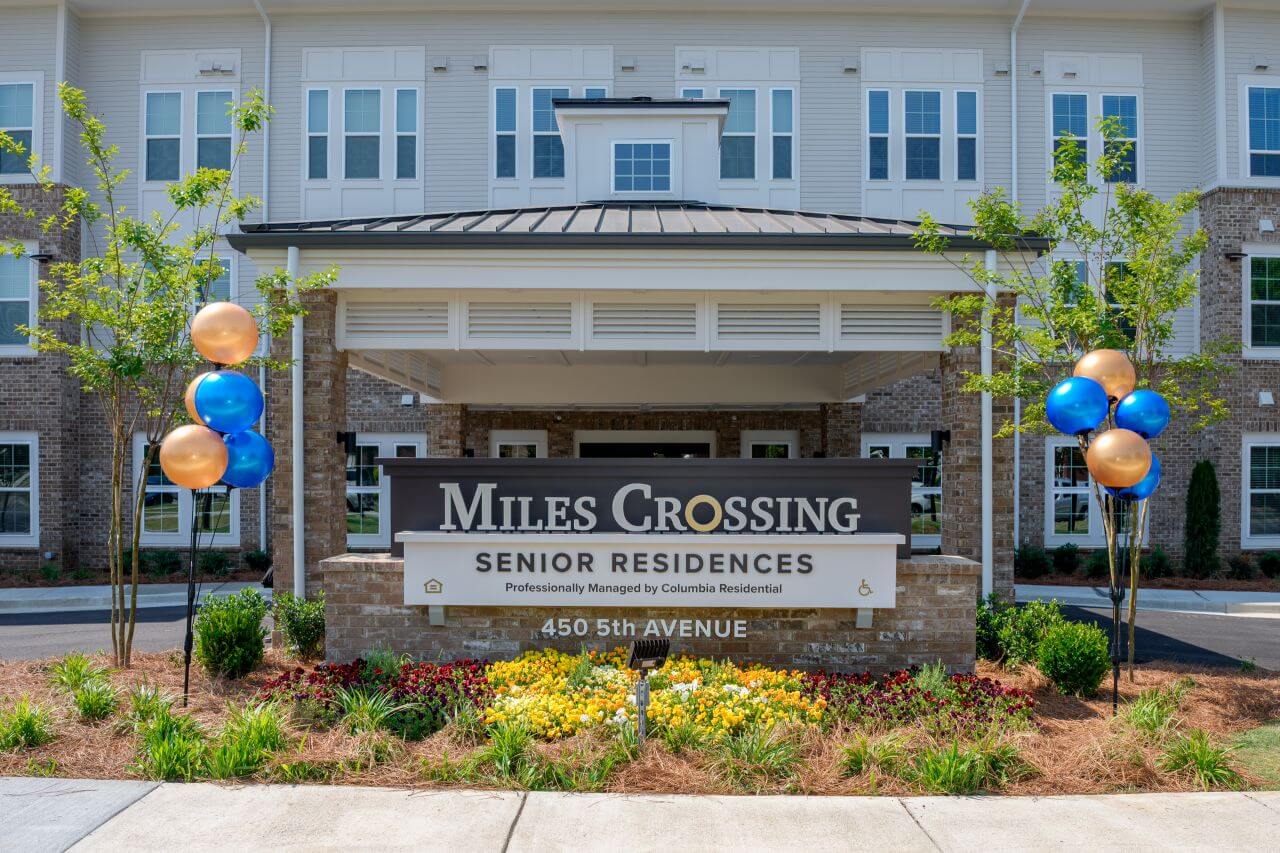 Entrance sign for Miles Crossing Senior Residences in front of a multi-story apartment building, with landscaped flowers and blue and gold balloons on either side, marking a celebratory event.