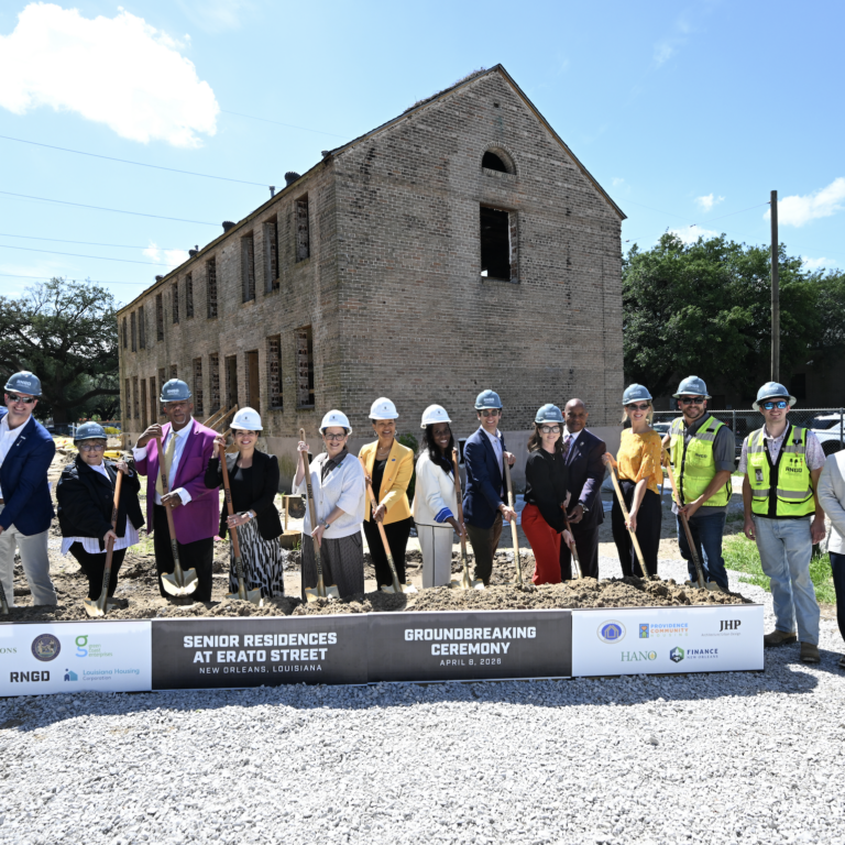 A group of project partners and community leaders wearing hard hats stand in front of a historic brick building, holding shovels during a groundbreaking ceremony for BW Cooper Senior. A sign in front reads “Senior Residences at Erato Street Groundbreaking Ceremony” in New Orleans, with construction underway on the site behind them.
