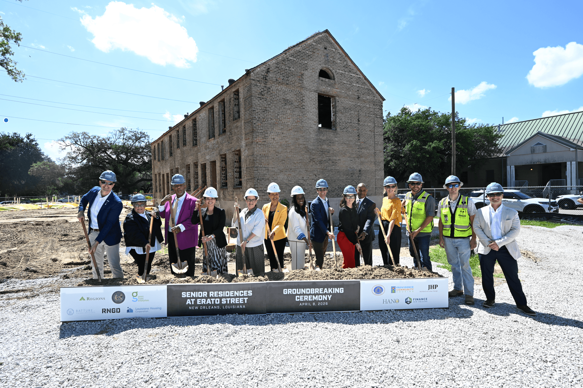 A group of project partners and community leaders wearing hard hats stand in front of a historic brick building, holding shovels during a groundbreaking ceremony for BW Cooper Senior. A sign in front reads “Senior Residences at Erato Street Groundbreaking Ceremony” in New Orleans, with construction underway on the site behind them.