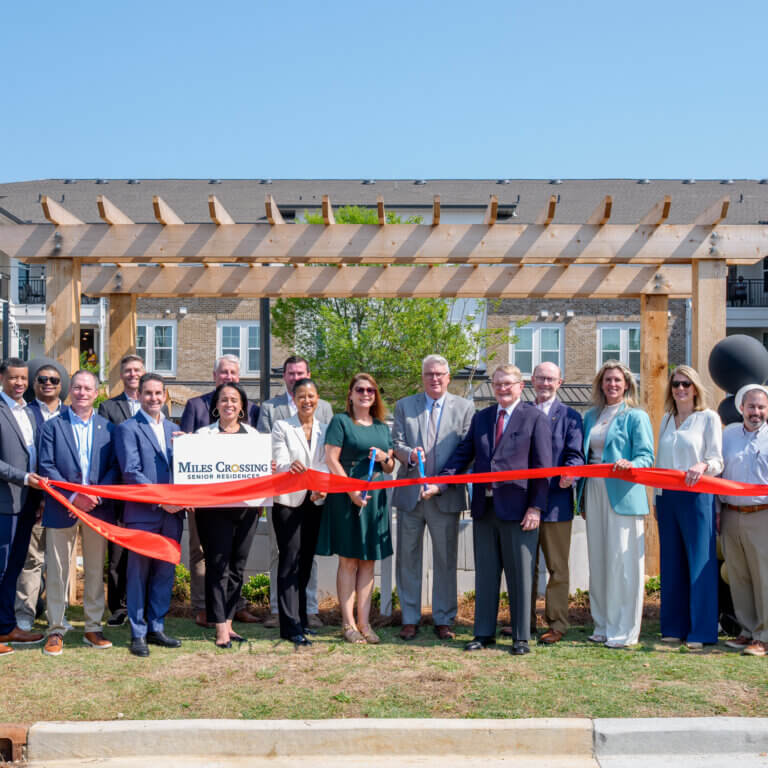 A group of community leaders, partners, and team members stand outdoors in front of a newly built apartment community, holding a red ribbon during a ribbon-cutting ceremony at Miles Crossing Senior Residences. A wooden pergola and multi-story residential buildings are visible in the background under a clear blue sky.