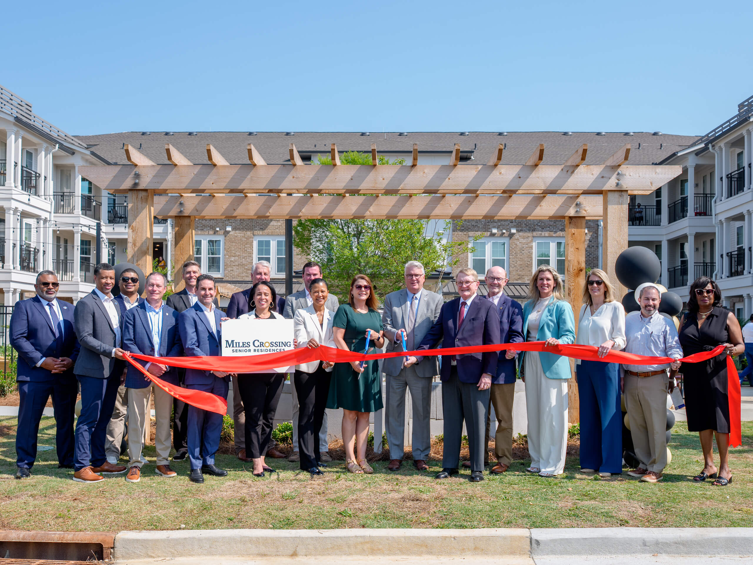 A group of community leaders, partners, and team members stand outdoors in front of a newly built apartment community, holding a red ribbon during a ribbon-cutting ceremony at Miles Crossing Senior Residences. A wooden pergola and multi-story residential buildings are visible in the background under a clear blue sky.