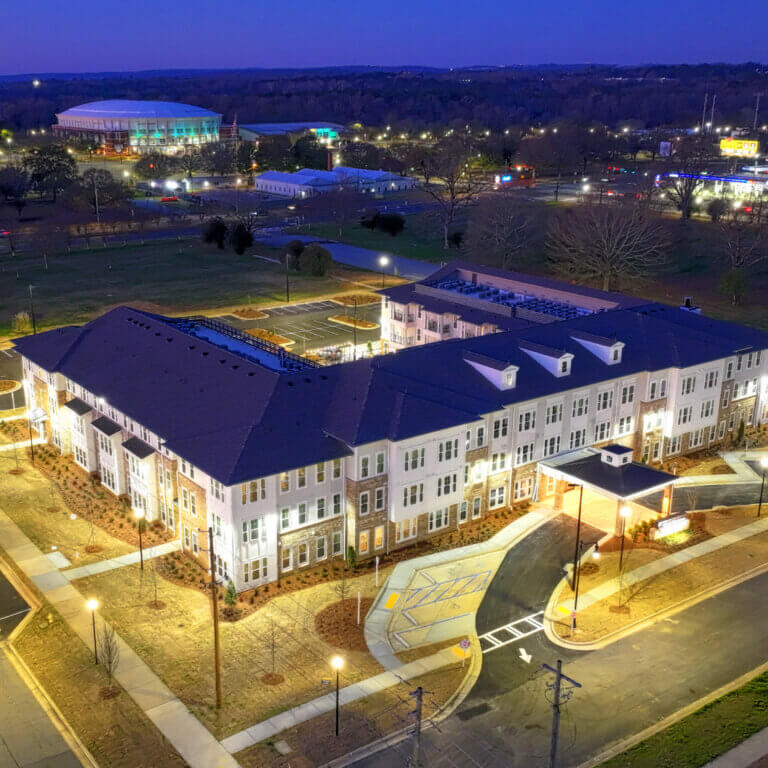 Aerial evening view of Miles Crossing Senior Residences, a multi-story apartment community with lit walkways, landscaped grounds, and a central entrance, surrounded by streets and nearby buildings. Miles Crossing brings more senior housing to Georgia.