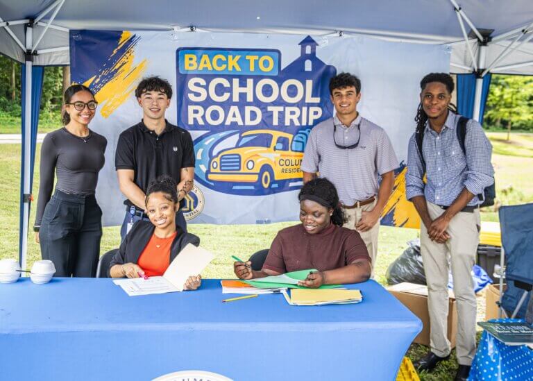 A group of young adults gather at a table during a Back to School Road Trip event, with a colorful school-themed banner in the background.