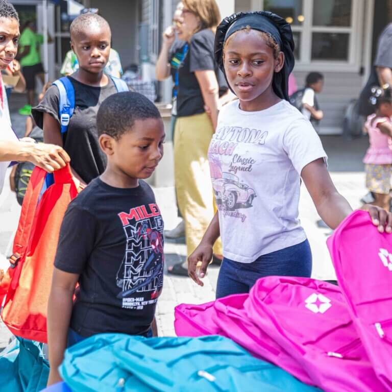 Two children stand outside looking at school supplies, including pink and blue backpacks. There are more people in the background.