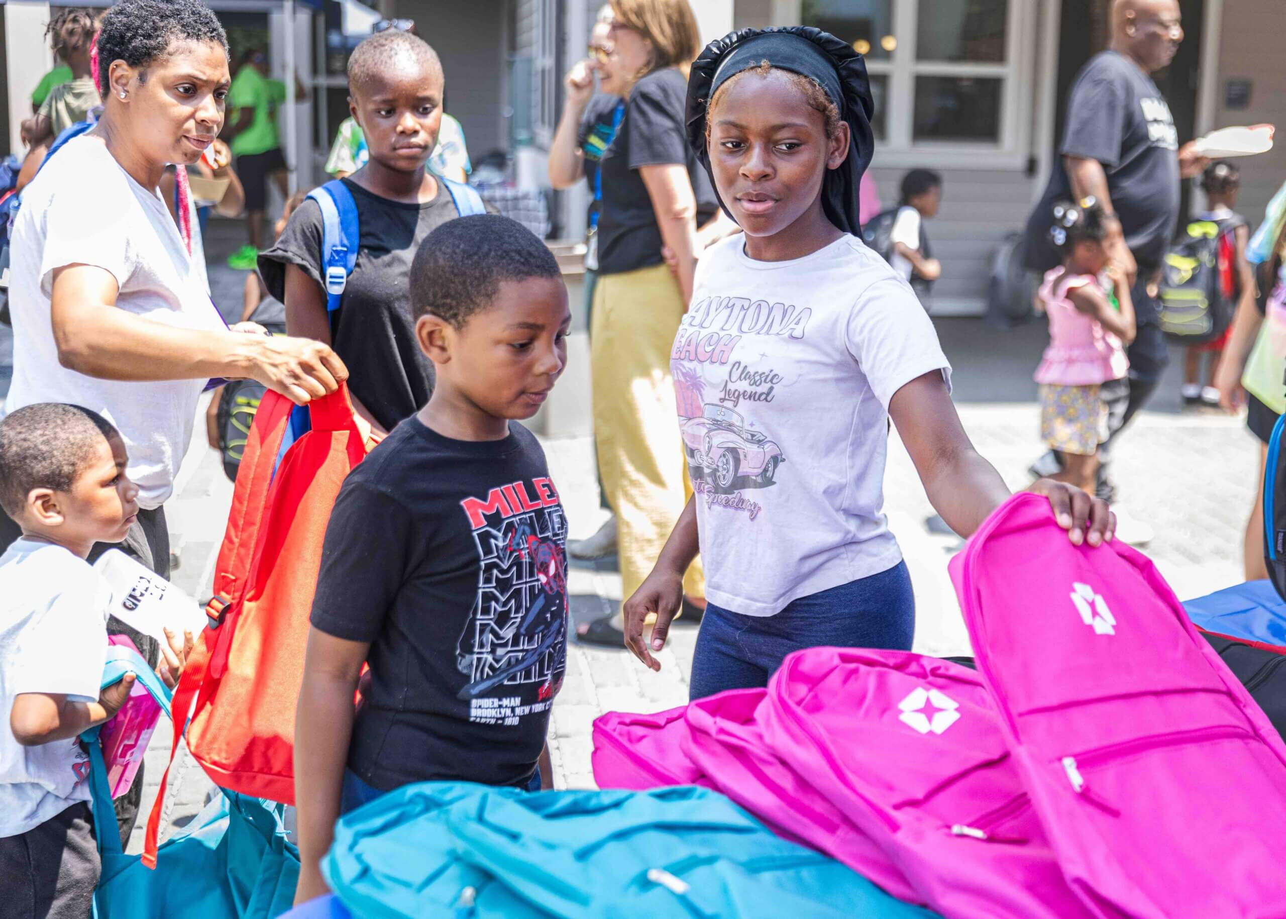 Two children stand outside looking at school supplies, including pink and blue backpacks. There are more people in the background.
