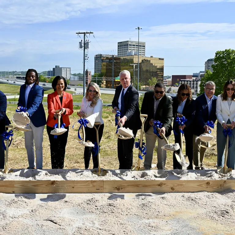 A diverse group of city leaders and community members breaking ground at a new development site in Chattanooga’s Westside, holding shovels with blue ribbon bows, against the backdrop of a city skyline and highway.