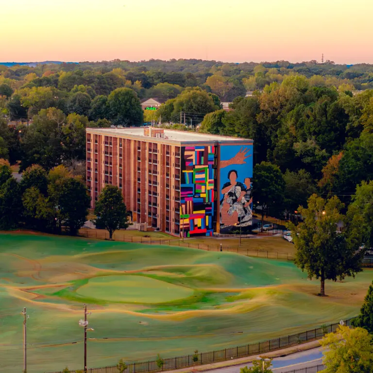 Aerial view of a mid-rise brick residential building featuring a large, colorful mural at Legacy at East Lake on its side, surrounded by trees and a nearby golf course under a warm sunset sky.