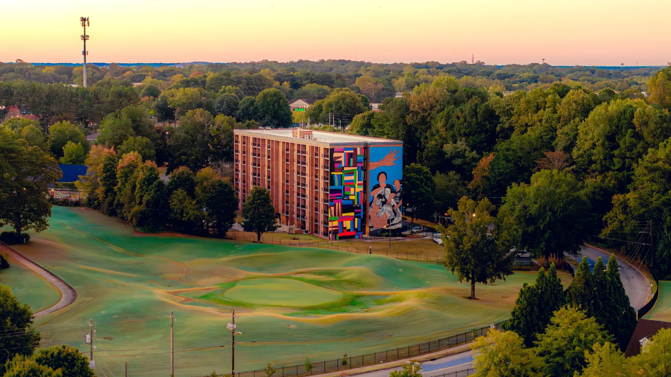 Aerial view of a mid-rise brick residential building featuring a large, colorful mural at Legacy at East Lake on its side, surrounded by trees and a nearby golf course under a warm sunset sky.