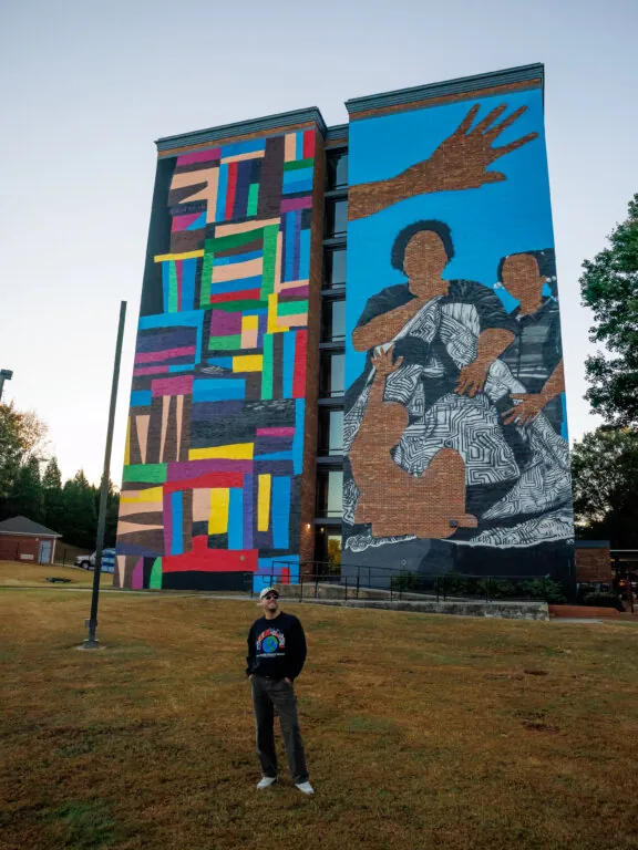 Artist Chastain ‘Chaz’ Clark stands in front of his large-scale mural on the side of a mid-rise residential building. The artwork features bold geometric color blocks on one side and stylized figures with an outstretched hand on a bright blue background on the other. 