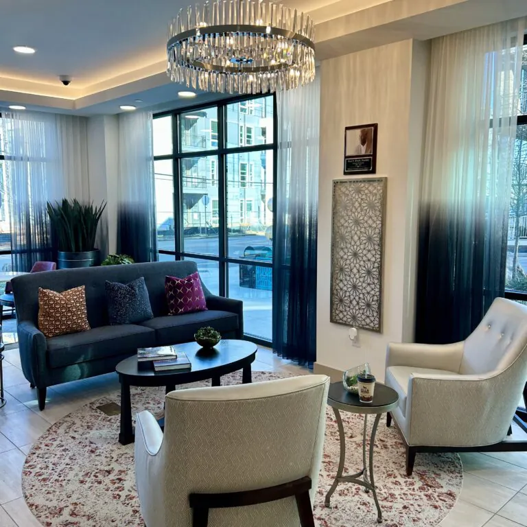 Modern lounge area at The Link featuring a gray sofa with accent pillows, two upholstered armchairs, a round coffee table, decorative rug, large windows with sheer curtains, and a contemporary chandelier overhead.