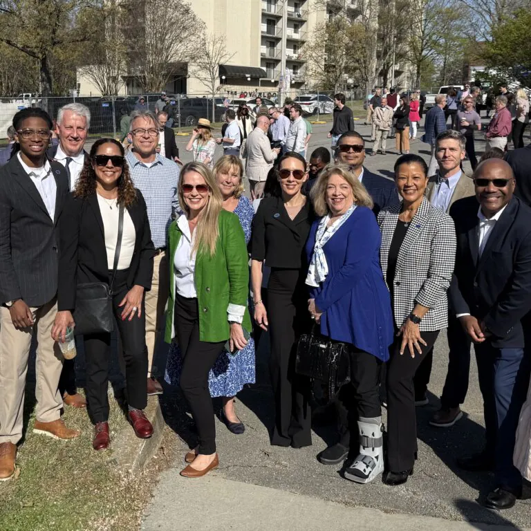 At the groundbreaking for Phase 2 of One Westside, a group of people standing outside, smiling at the camera. Trees, building, and a construction fence are visible in the background.