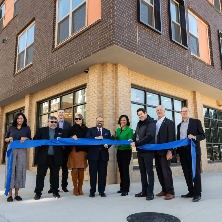 A group of community leaders and project partners stand outside the newly constructed apartment building at a senior housing development in Decatur — The Link. These individuals are holding a long blue ribbon during a ribbon-cutting ceremony. The modern mixed-use building features brick and large windows, with ground-floor retail space and residential units above, marking the opening of a new housing community.