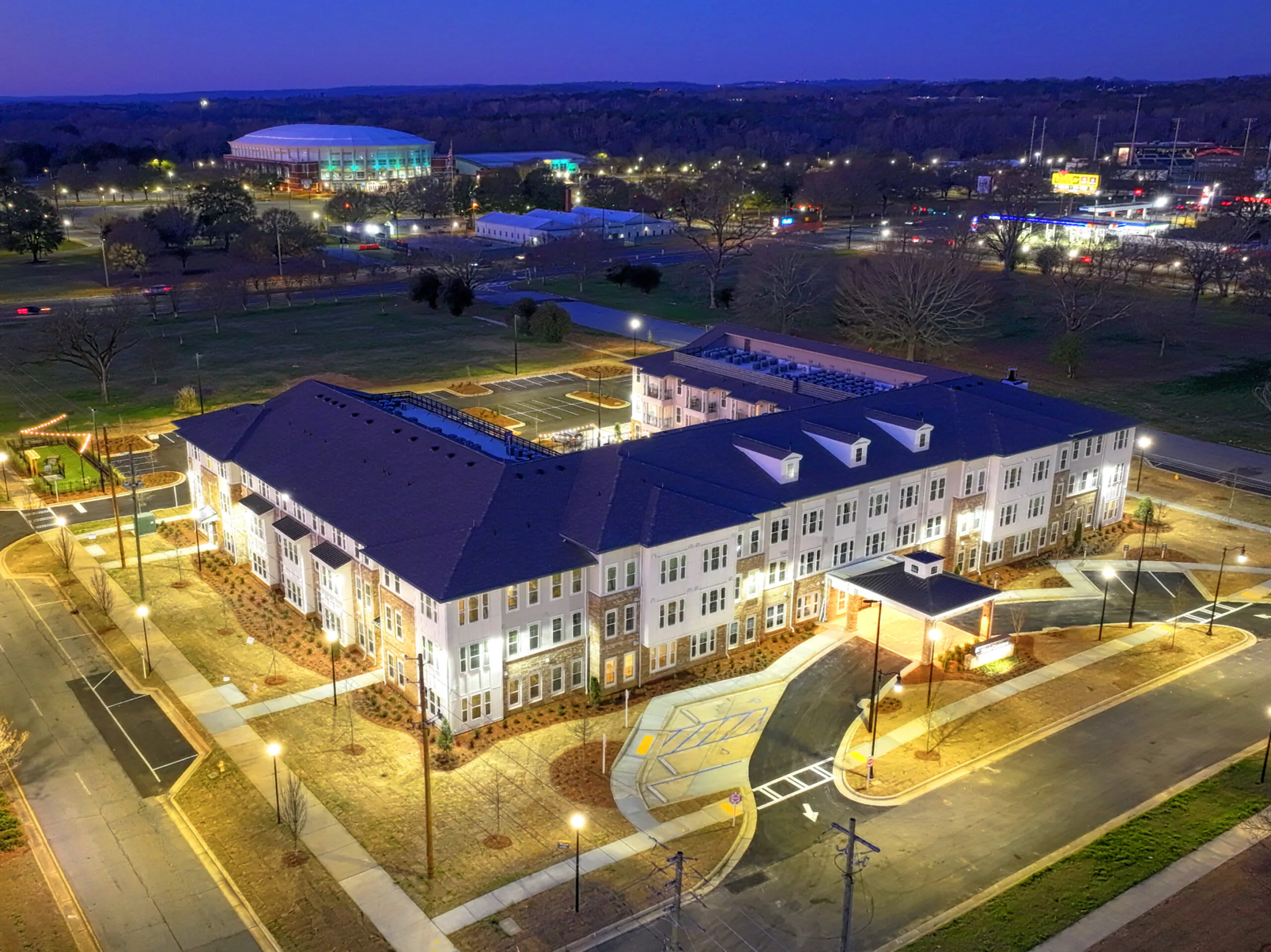 Aerial evening view of Miles Crossing Senior Residences, a multi-story apartment community with lit walkways, landscaped grounds, and a central entrance, surrounded by streets and nearby buildings. Miles Crossing brings more senior housing to Georgia.